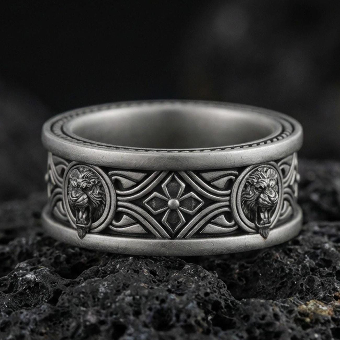Oxidized sterling silver lion medallion band ring on dark volcanic rock, twin lion head relief panels with gothic cross and scrollwork center, premium macro product shot.