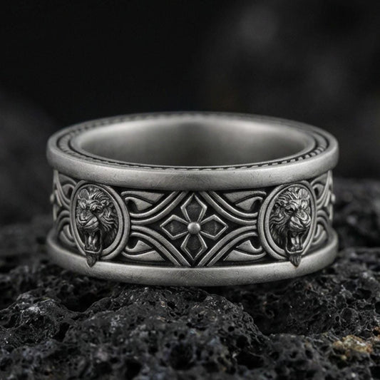 Oxidized sterling silver lion medallion band ring on dark volcanic rock, twin lion head relief panels with gothic cross and scrollwork center, premium macro product shot.