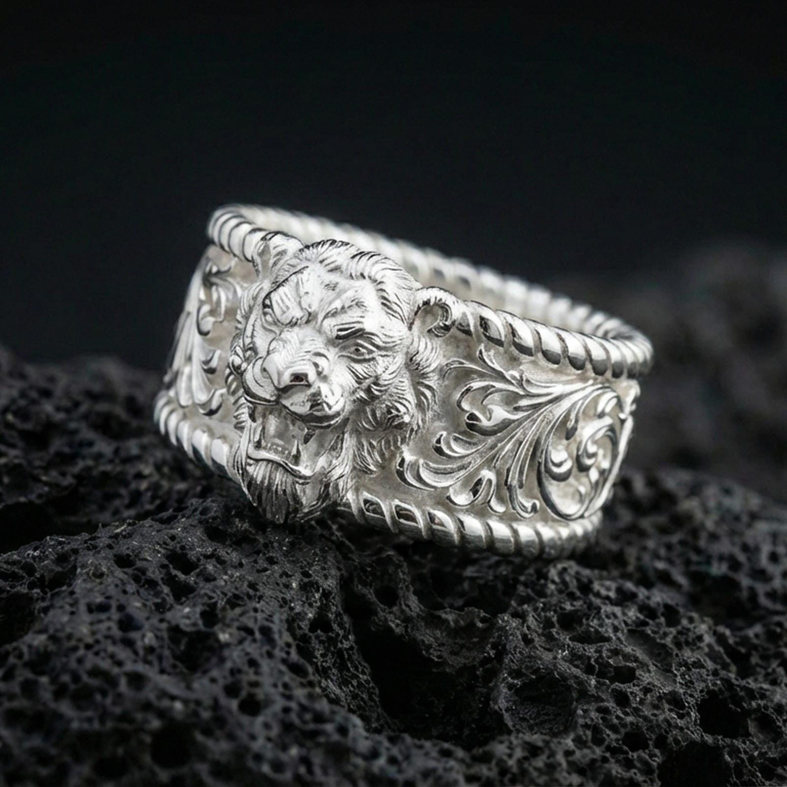 Polished silver lion signet ring on black volcanic stone, crisp lion head relief with detailed mane and engraved scrollwork sides, centered close-up studio lighting.
