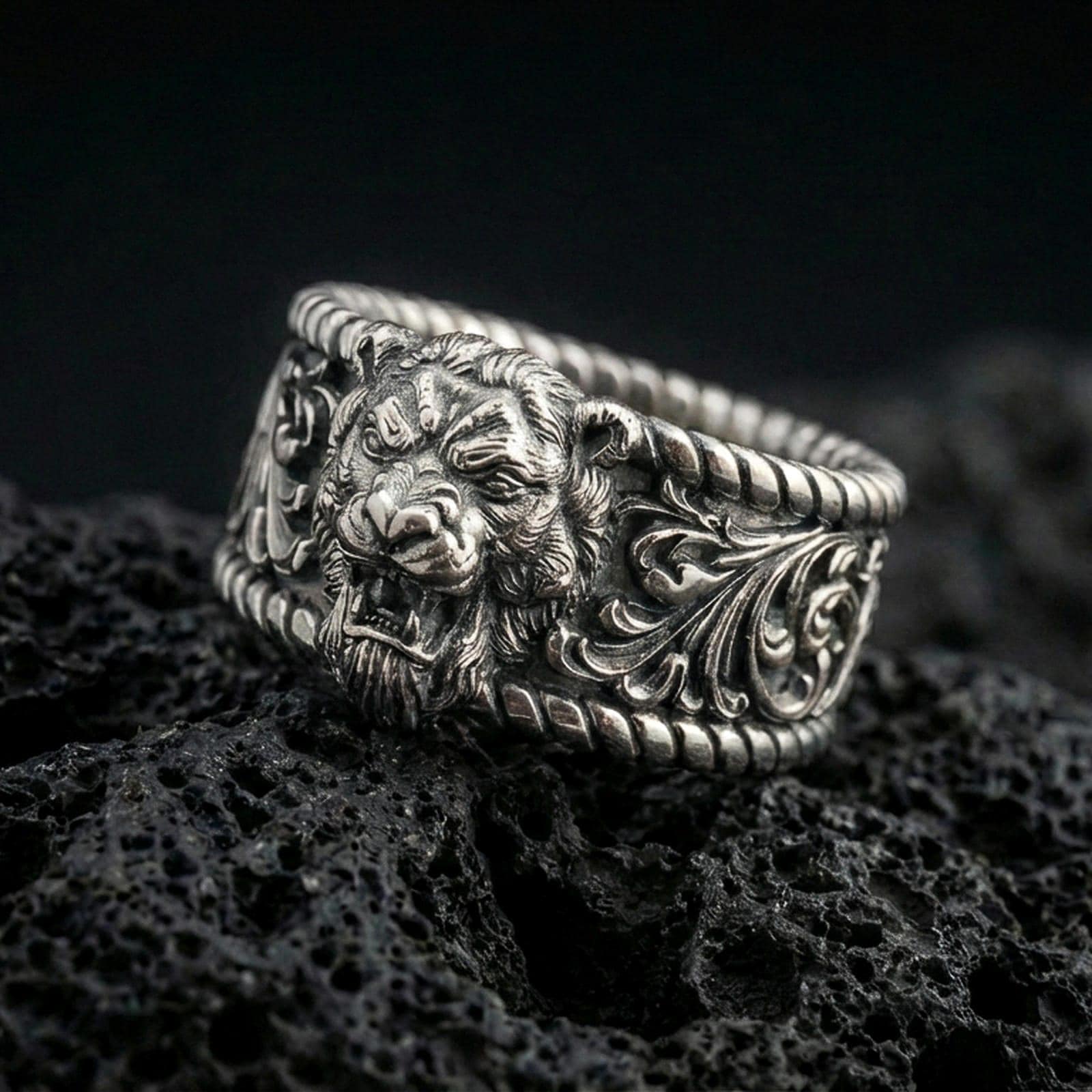 Oxidized sterling silver lion head signet ring on dark volcanic rock, bold roaring lion relief with ornate scrollwork shoulders and rope-edge borders, premium macro product shot.