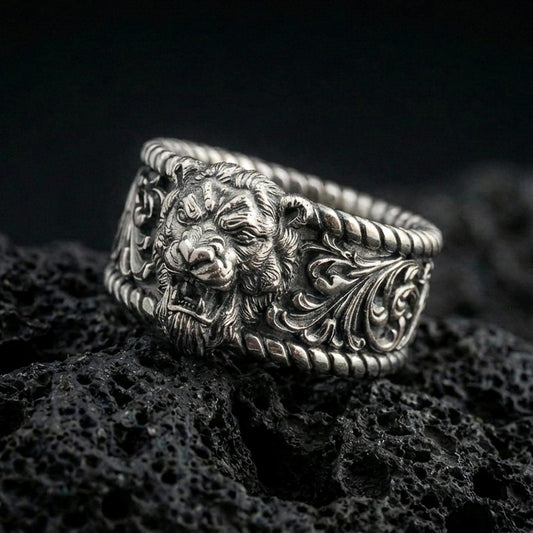 Oxidized sterling silver lion head signet ring on dark volcanic rock, bold roaring lion relief with ornate scrollwork shoulders and rope-edge borders, premium macro product shot.