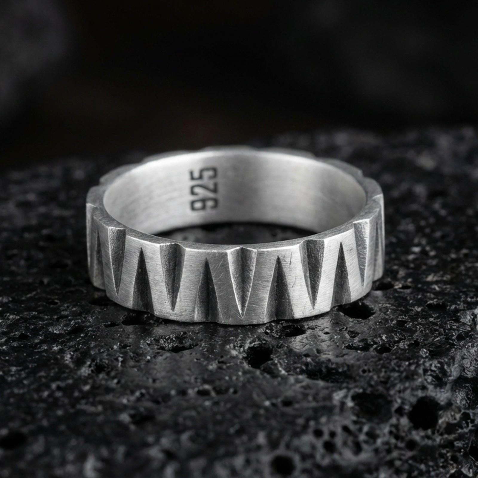 Oxidized sterling silver band ring on dark volcanic rock, bold faceted chevron cuts with deep shadowed grooves, clean macro product photography.
