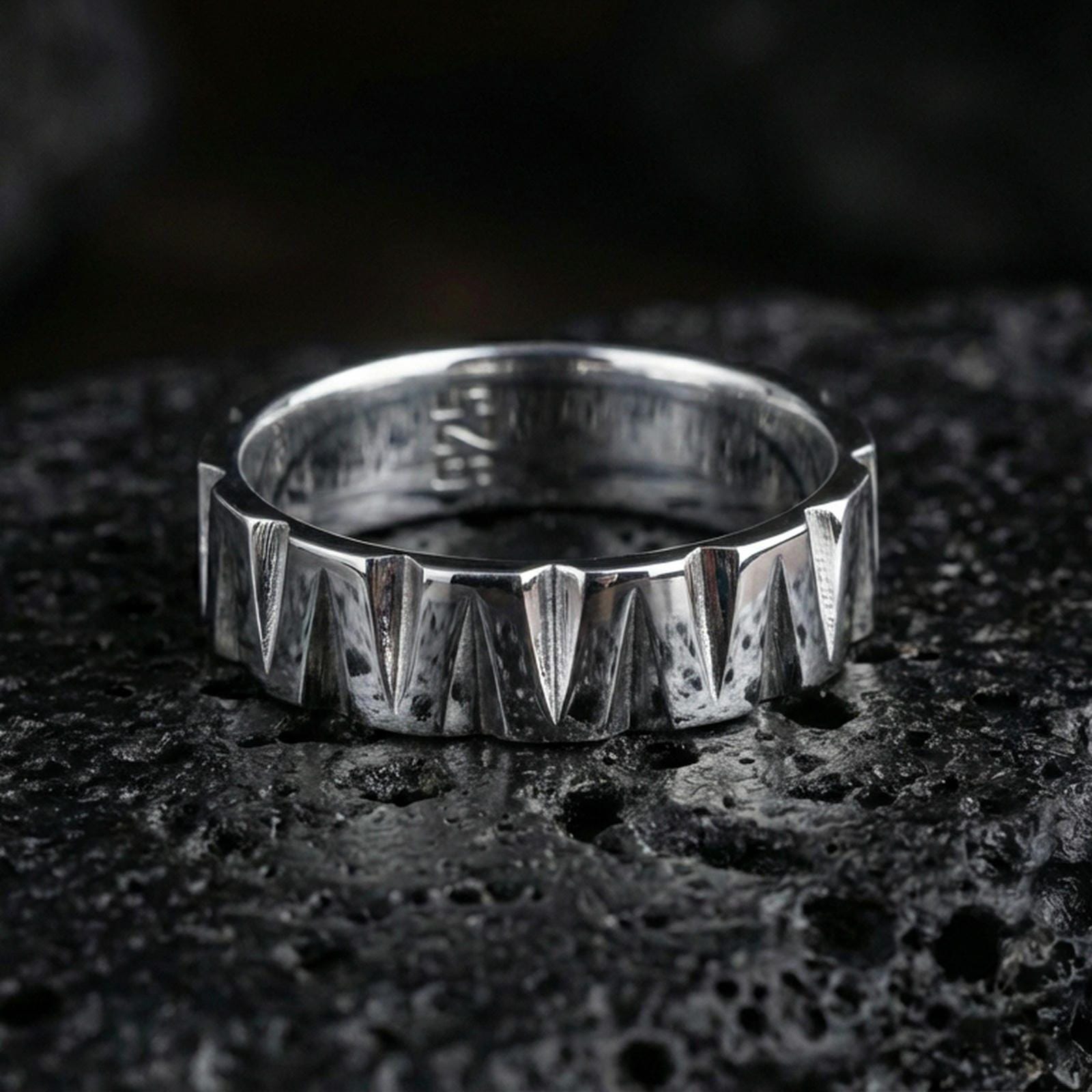 Polished silver geometric band ring on black volcanic stone, crisp angular wedge facets with bright highlights and brushed texture, centered close-up.