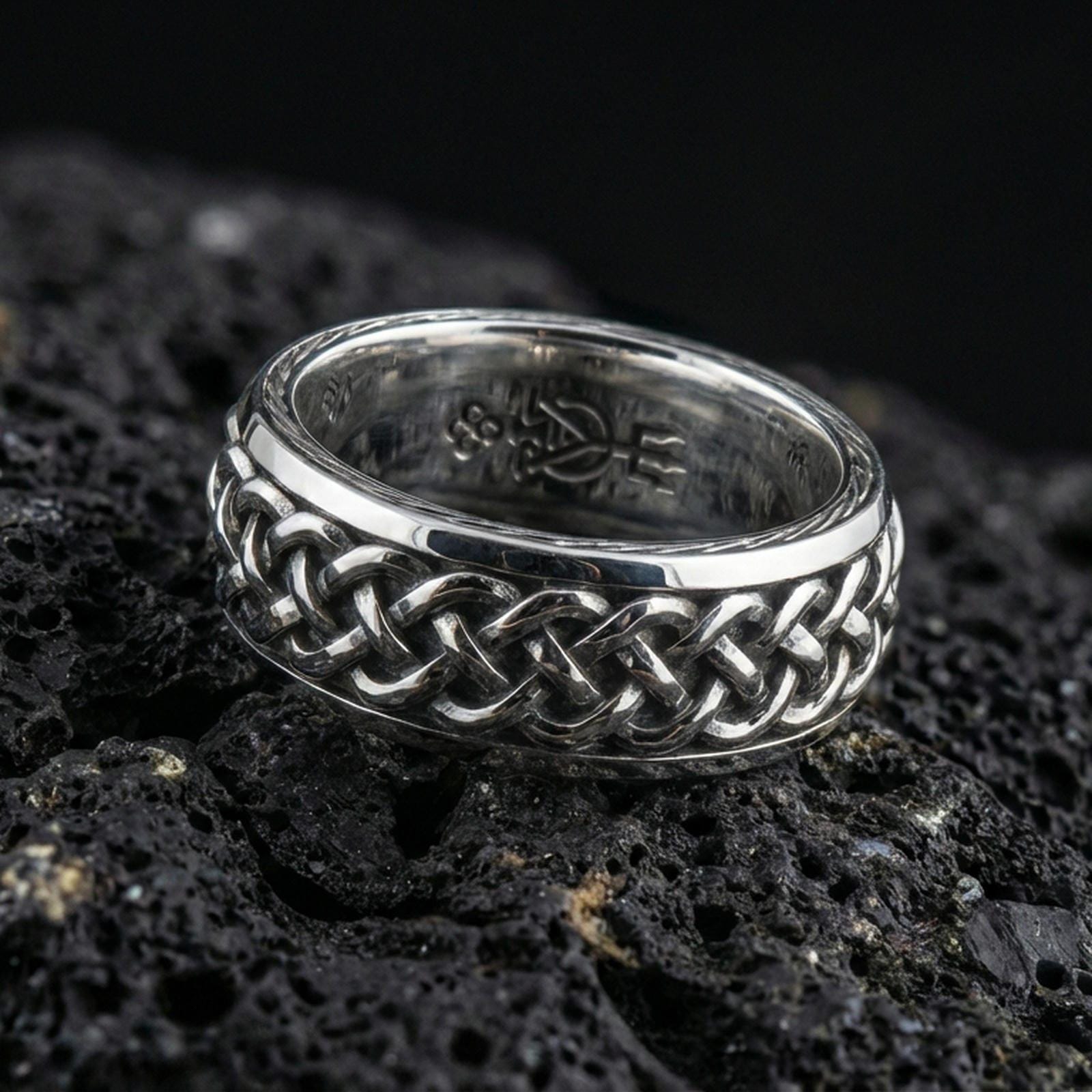 Polished silver braided Celtic knot ring on dark volcanic stone, bright reflective weave links with clean edges, premium close-up jewelry photography.
