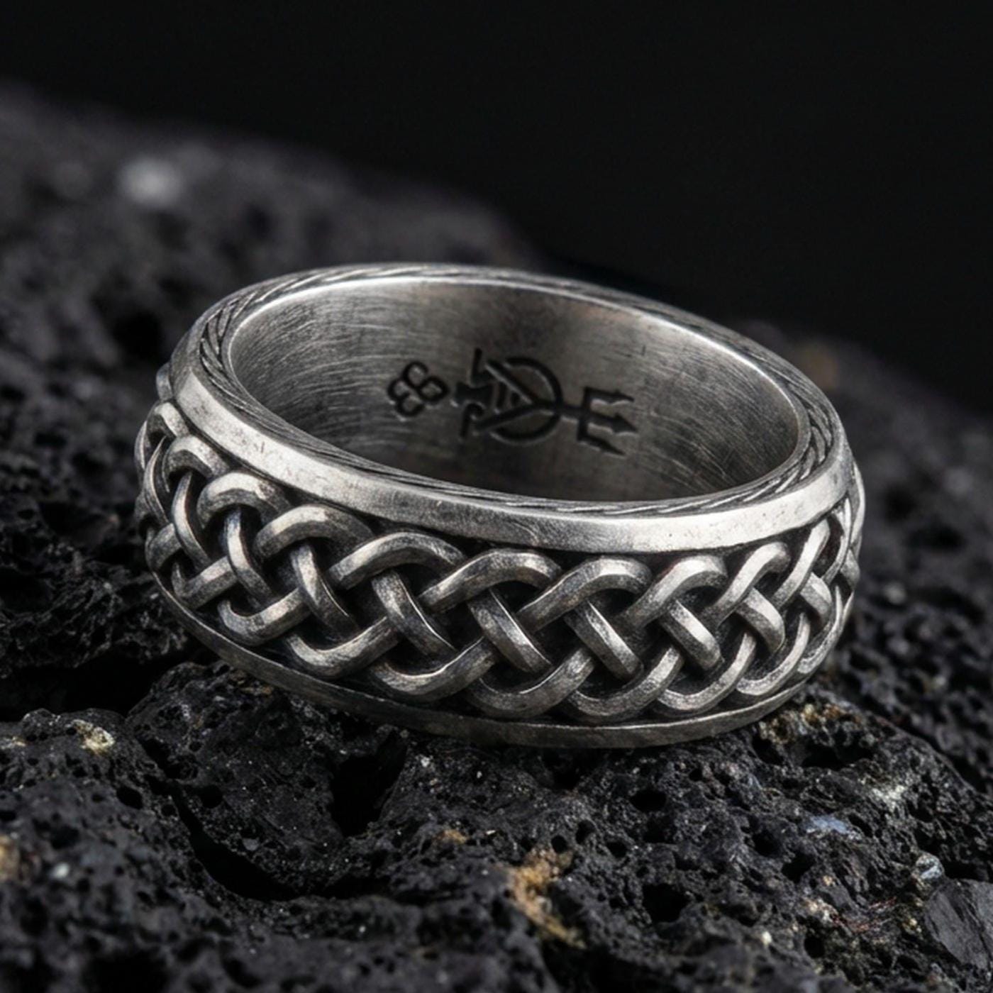 Oxidized sterling silver Celtic knot band ring on black volcanic rock, raised interwoven braid pattern with deep shadowed recesses, crisp macro product shot.