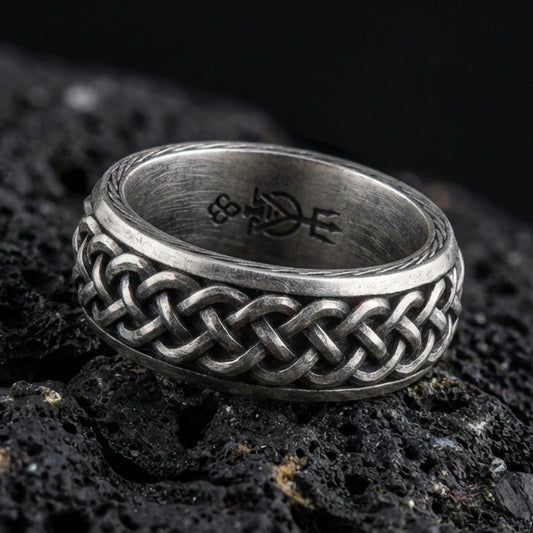 Oxidized sterling silver Celtic knot band ring on black volcanic rock, raised interwoven braid pattern with deep shadowed recesses, crisp macro product shot.