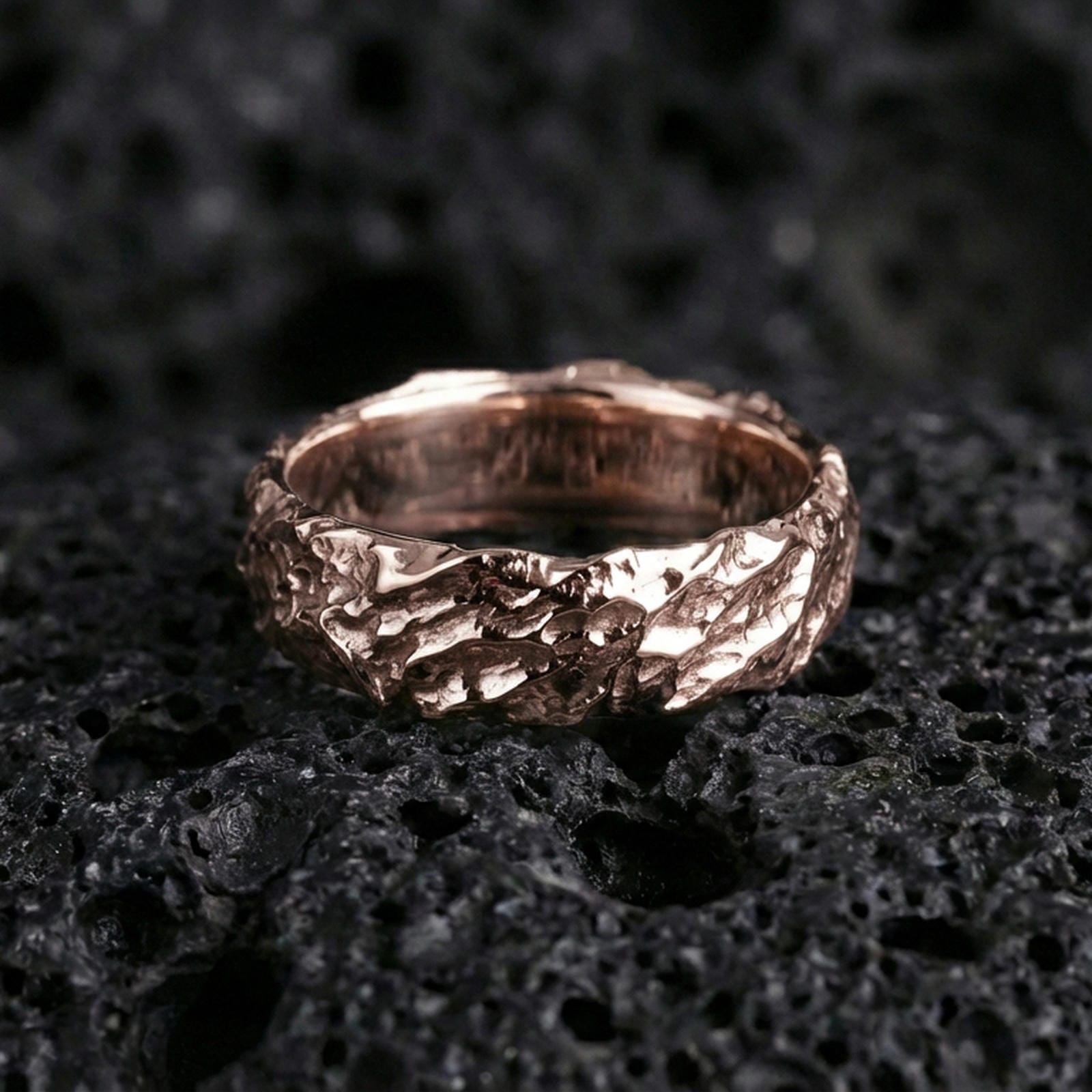 Rose gold plated hammered texture band on black volcanic stone, organic rocky relief with soft warm shine, luxury product photo.