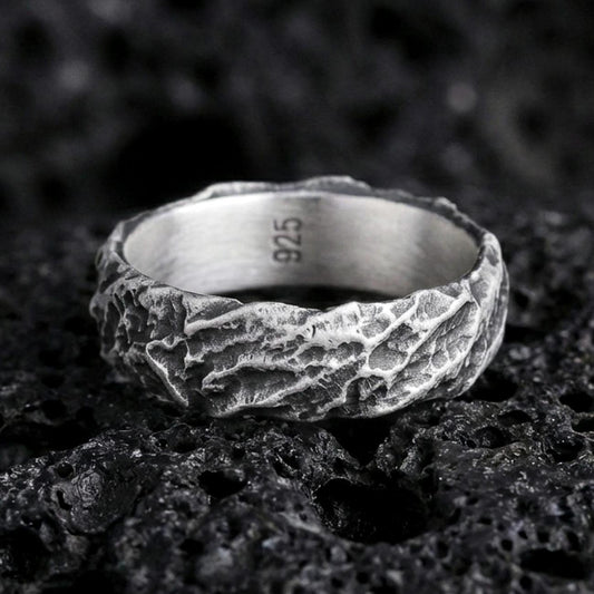 Oxidized sterling silver hammered texture band ring on black volcanic rock, rugged molten-stone surface with deep shadowed crevices, macro product photo.