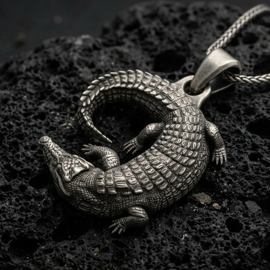 Oxidized crocodile pendant with detailed reptile scales, curved tail, and realistic animal silhouette displayed on dark volcanic rock in a close-up luxury jewelry composition