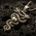 Load image into Gallery viewer, Bronze serpent pendant with coiled snake, apple detail, and branch motif displayed on dark volcanic rock in a close-up luxury jewelry composition
