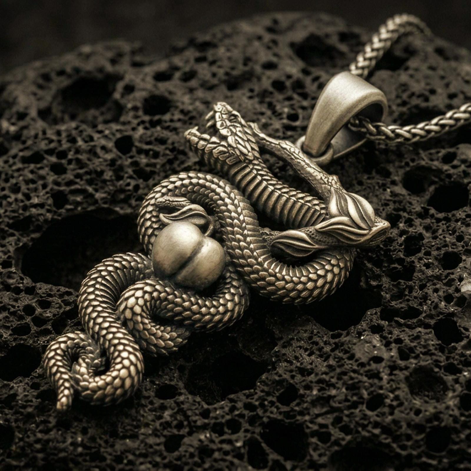 Bronze serpent pendant with coiled snake, apple detail, and branch motif displayed on dark volcanic rock in a close-up luxury jewelry composition