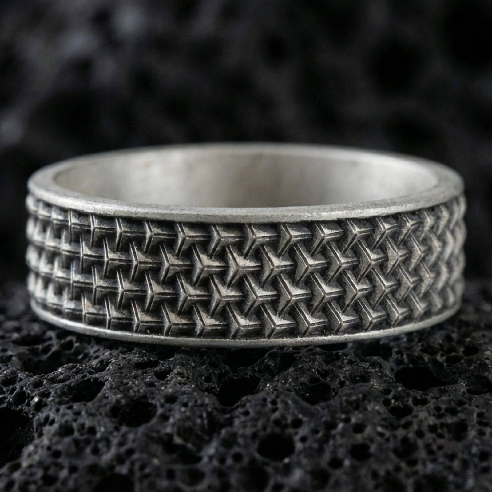 Oxidized sterling silver woven-grid texture band ring on black volcanic rock, deep shadowed recesses emphasizing the geometric pattern, macro product photo.