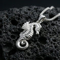 Load image into Gallery viewer, Polished silver seahorse necklace with detailed ridged body, curled tail, and dimensional ocean-inspired form placed on textured volcanic rock in a premium studio product shot
