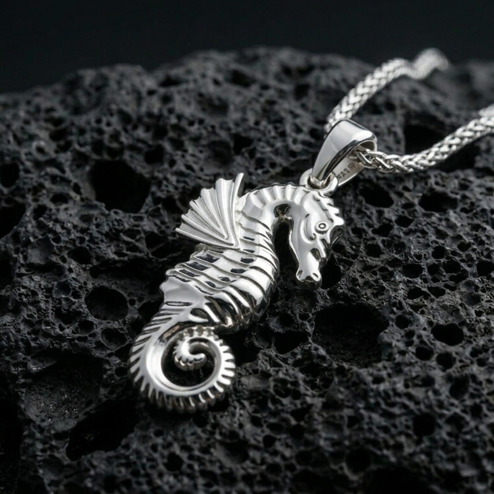 Polished silver seahorse necklace with detailed ridged body, curled tail, and dimensional ocean-inspired form placed on textured volcanic rock in a premium studio product shot