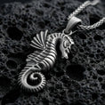 Load image into Gallery viewer, Oxidized seahorse pendant with sculpted marine details, curled tail, and textured nautical silhouette displayed on dark volcanic rock in a close-up luxury jewelry composition
