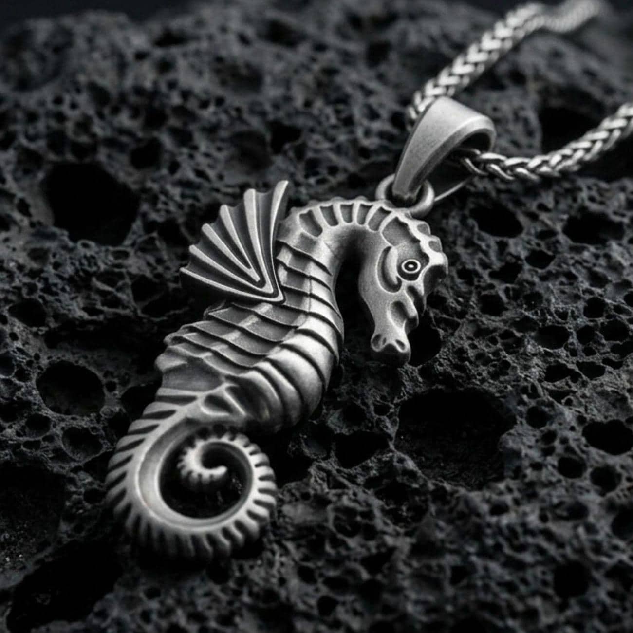 Oxidized seahorse pendant with sculpted marine details, curled tail, and textured nautical silhouette displayed on dark volcanic rock in a close-up luxury jewelry composition