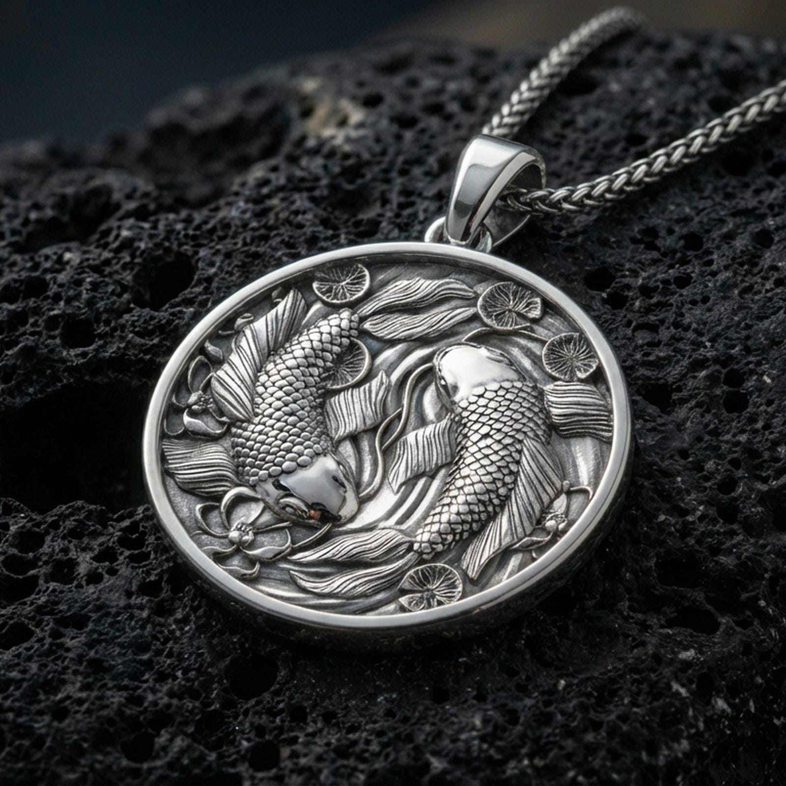 Polished silver koi necklace with detailed fish scales, circular yin yang arrangement, and layered aquatic floral motifs placed on textured volcanic rock in a premium studio product shot