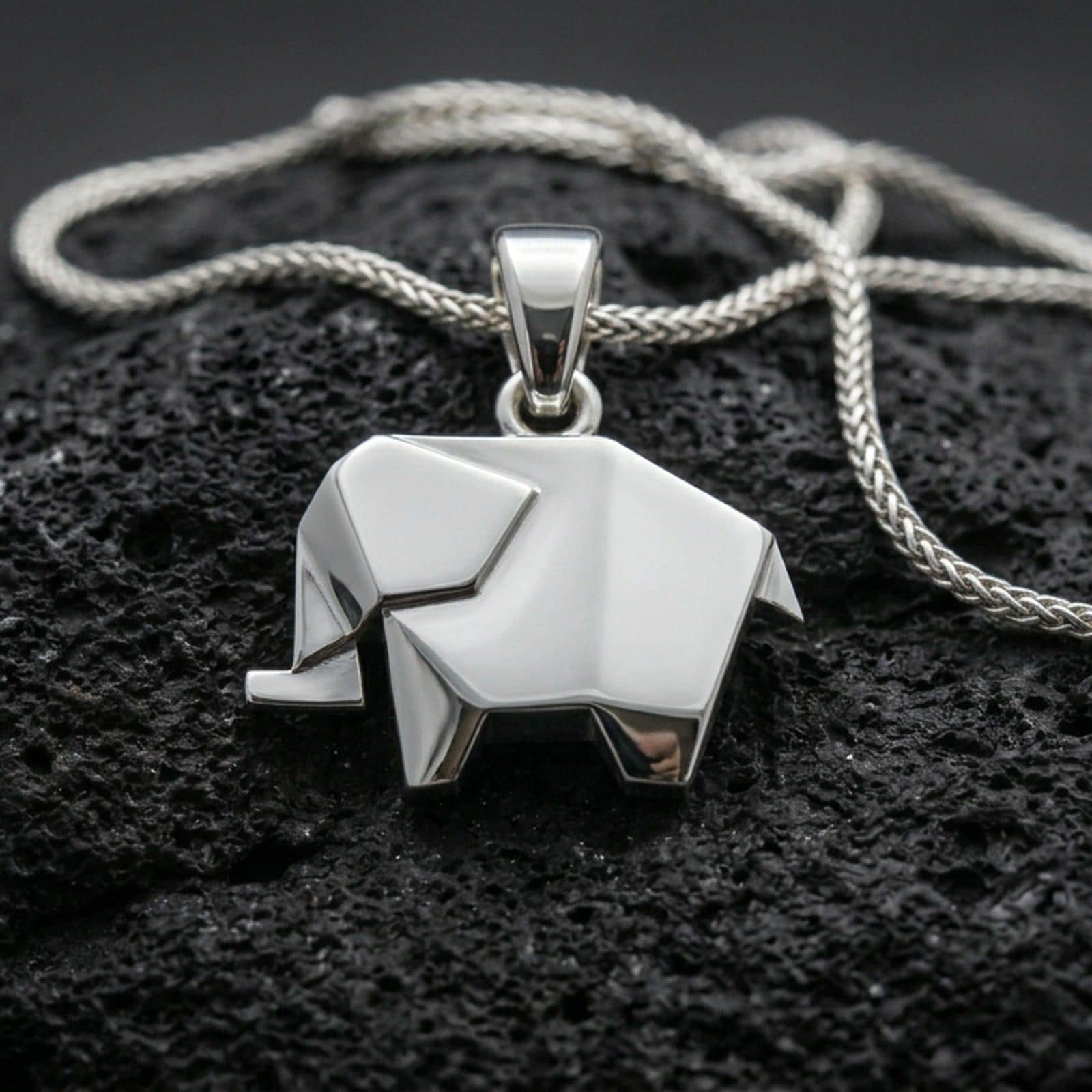 Polished silver elephant necklace with angular origami-inspired structure, clean faceted surfaces, and modern animal design placed on textured volcanic rock in a premium studio product shot