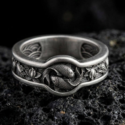 Oxidized sterling silver Yin Yang koi fish band ring on black volcanic stone, darkened relief emphasizing the twin koi and lotus side details, macro product photo.