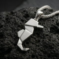 Load image into Gallery viewer, Polished silver seahorse necklace with angular origami-inspired structure, faceted folded surfaces, and sleek nautical form placed on textured volcanic rock in a premium studio product shot
