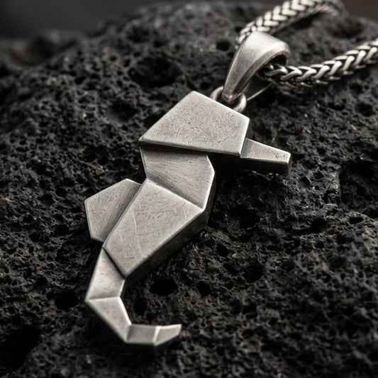 Oxidized origami seahorse pendant with geometric folded lines and minimalist marine silhouette displayed on dark volcanic rock in a close-up luxury jewelry composition