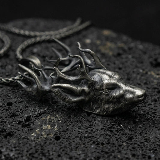 Oxidized stag pendant with detailed deer head, branching antlers, and realistic wildlife silhouette displayed on dark volcanic rock in a close-up luxury jewelry composition