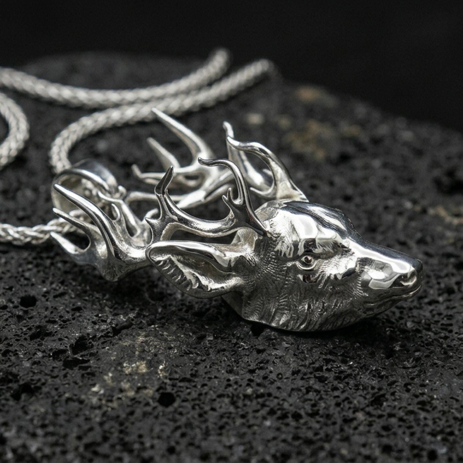 Polished silver deer necklace with sculpted stag face, dramatic antler spread, and lifelike animal form placed on textured volcanic rock in a premium studio product shot