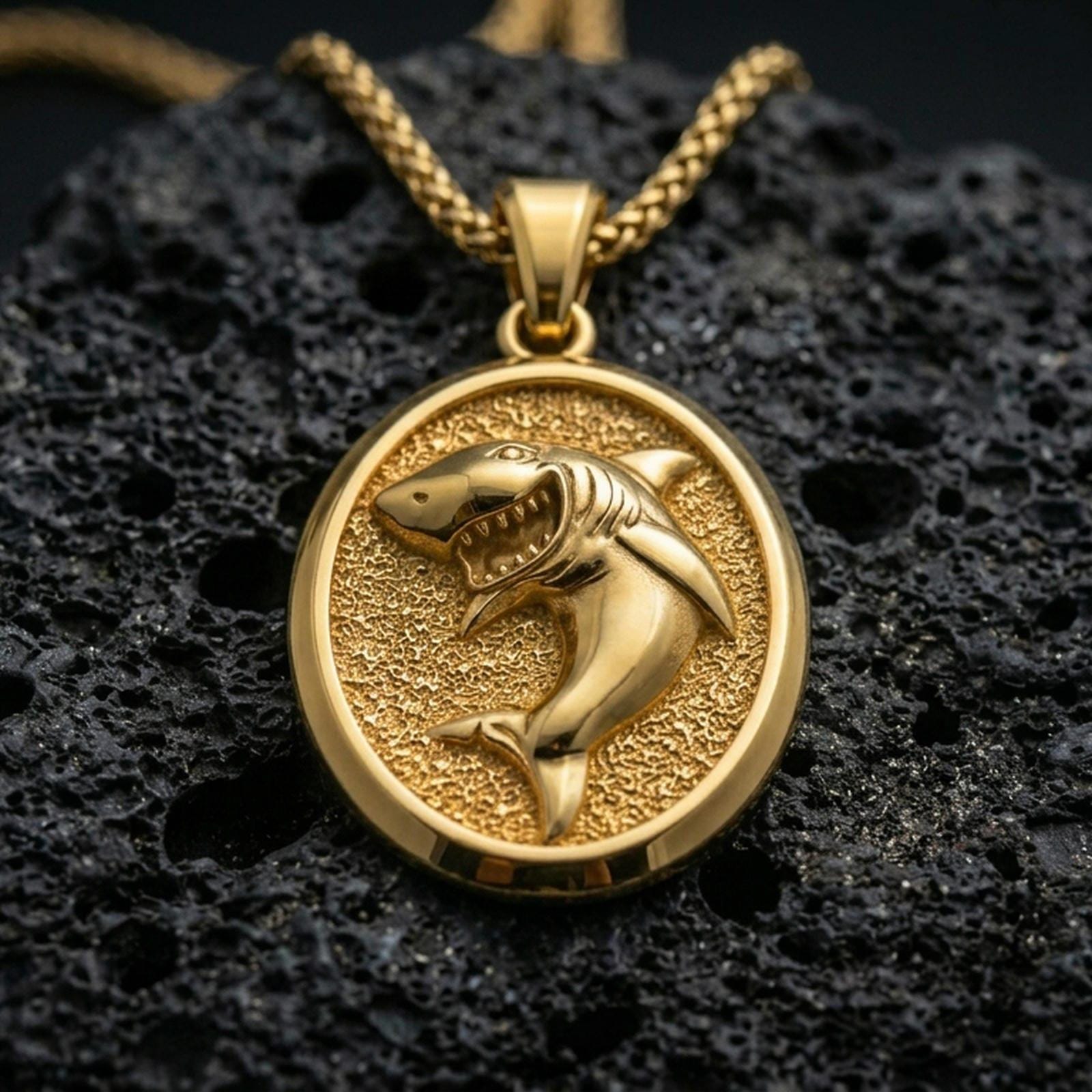 Gold plated shark pendant with bold shark figure, open jaws, and classic oval medallion form showcased on black volcanic rock in a refined macro jewelry image
