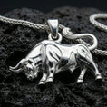 Load image into Gallery viewer, Polished silver bull necklace with sculpted body contours, detailed horn features, and bold animal silhouette placed on textured volcanic rock in a premium studio product shot
