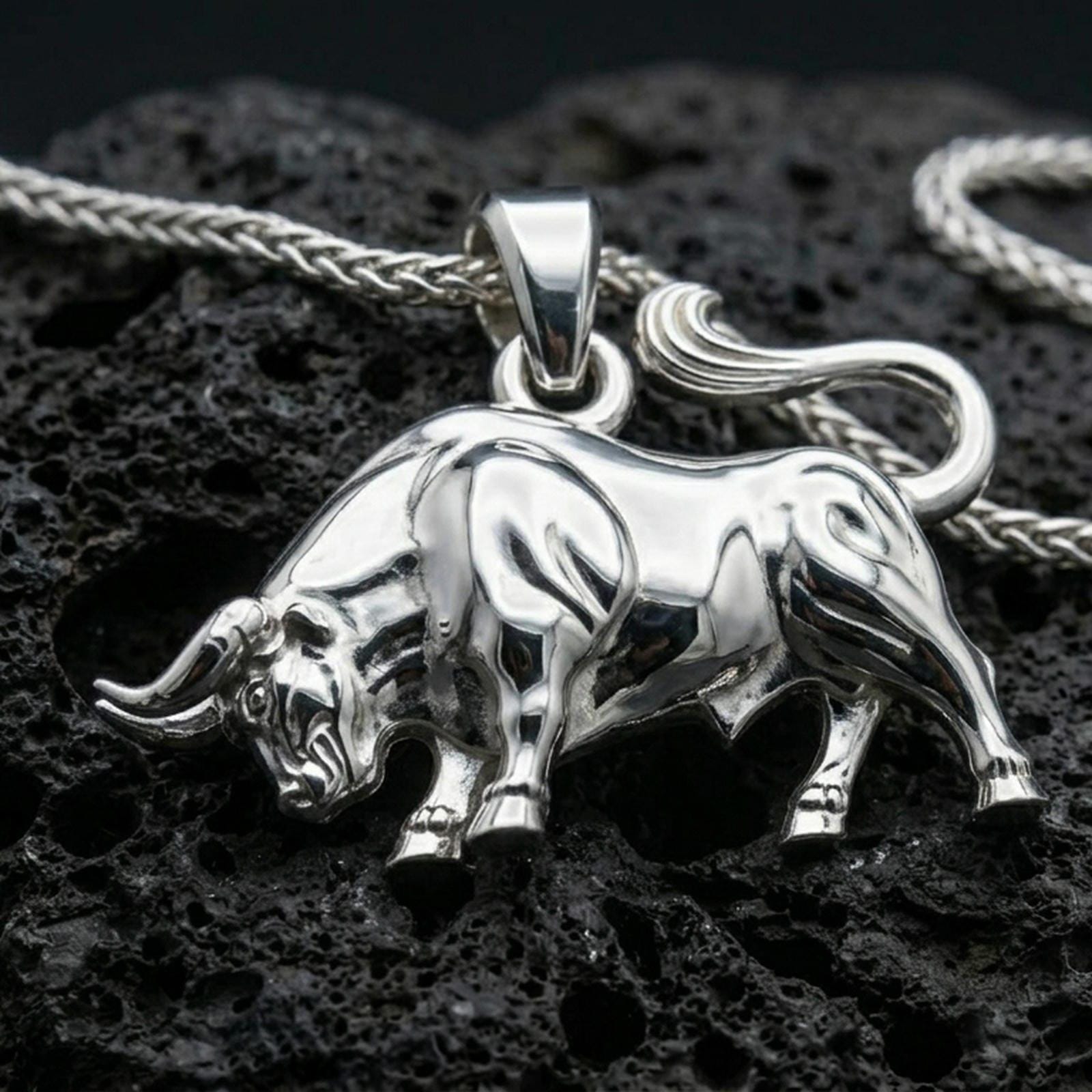 Polished silver bull necklace with sculpted body contours, detailed horn features, and bold animal silhouette placed on textured volcanic rock in a premium studio product shot