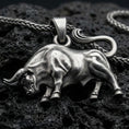 Load image into Gallery viewer, Oxidized bull pendant with muscular animal form, curved tail, and strong charging stance displayed on dark volcanic rock in a close-up luxury jewelry composition
