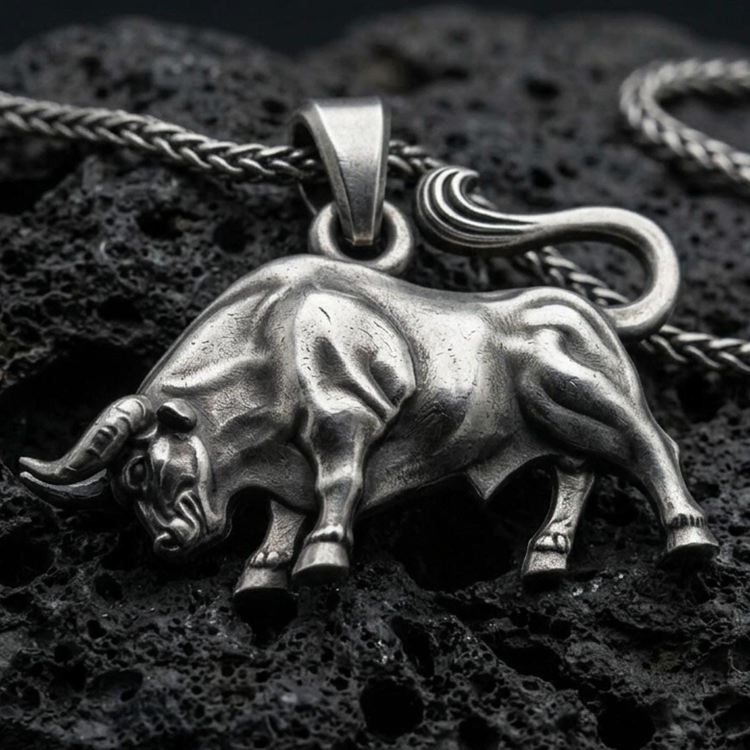 Oxidized bull pendant with muscular animal form, curved tail, and strong charging stance displayed on dark volcanic rock in a close-up luxury jewelry composition