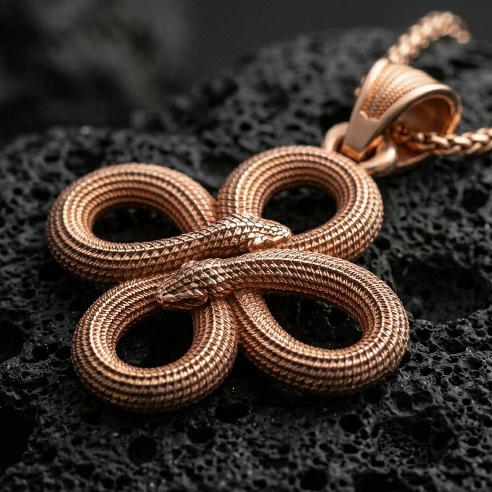 Rose gold plated serpent pendant with intertwined snake design, detailed coiled texture, and balanced symbolic knot form resting on volcanic rock in an elegant close-up product presentation