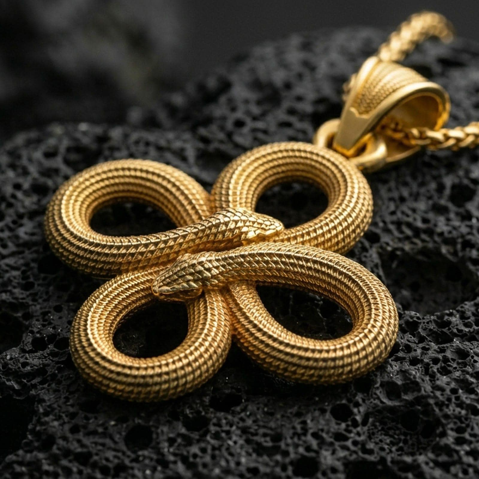 Gold plated snake knot pendant with interwoven serpent bodies, realistic scale relief, and elegant endless loop silhouette showcased on black volcanic rock in a refined macro jewelry image