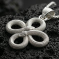 Load image into Gallery viewer, Polished silver serpent necklace with twin snake heads, intricate coiled knot form, and textured scale detailing placed on textured volcanic rock in a premium studio product shot
