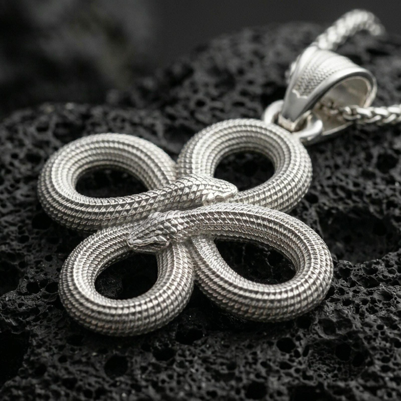 Polished silver serpent necklace with twin snake heads, intricate coiled knot form, and textured scale detailing placed on textured volcanic rock in a premium studio product shot
