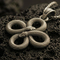 Load image into Gallery viewer, Bronze snake knot pendant with intertwined serpents, detailed scale texture, and symmetrical looped design displayed on dark volcanic rock in a close-up luxury jewelry composition
