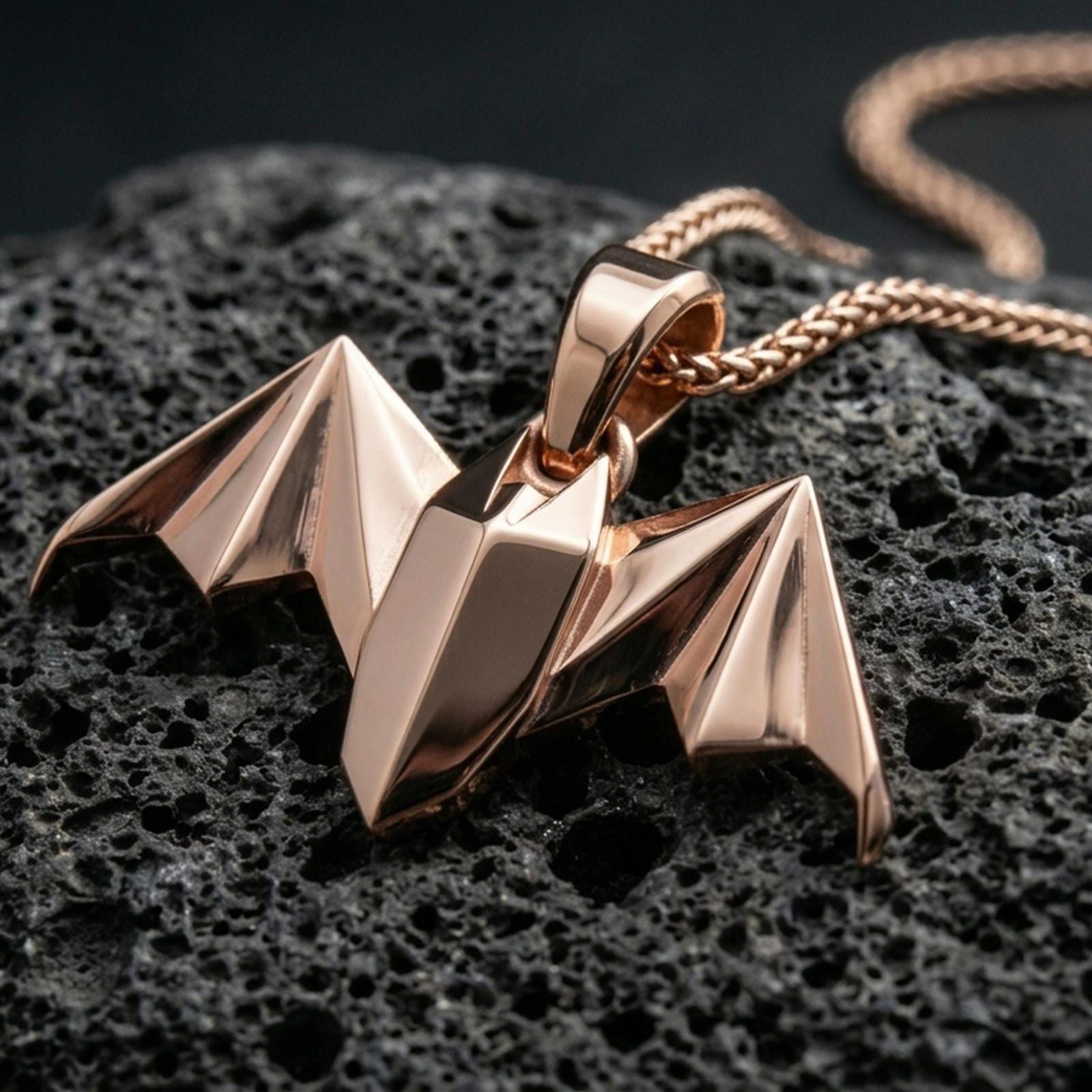 Rose gold plated bat pendant with angular origami wings, minimalist gothic form, and elegant sculptural silhouette resting on volcanic rock in an elegant close-up product presentation