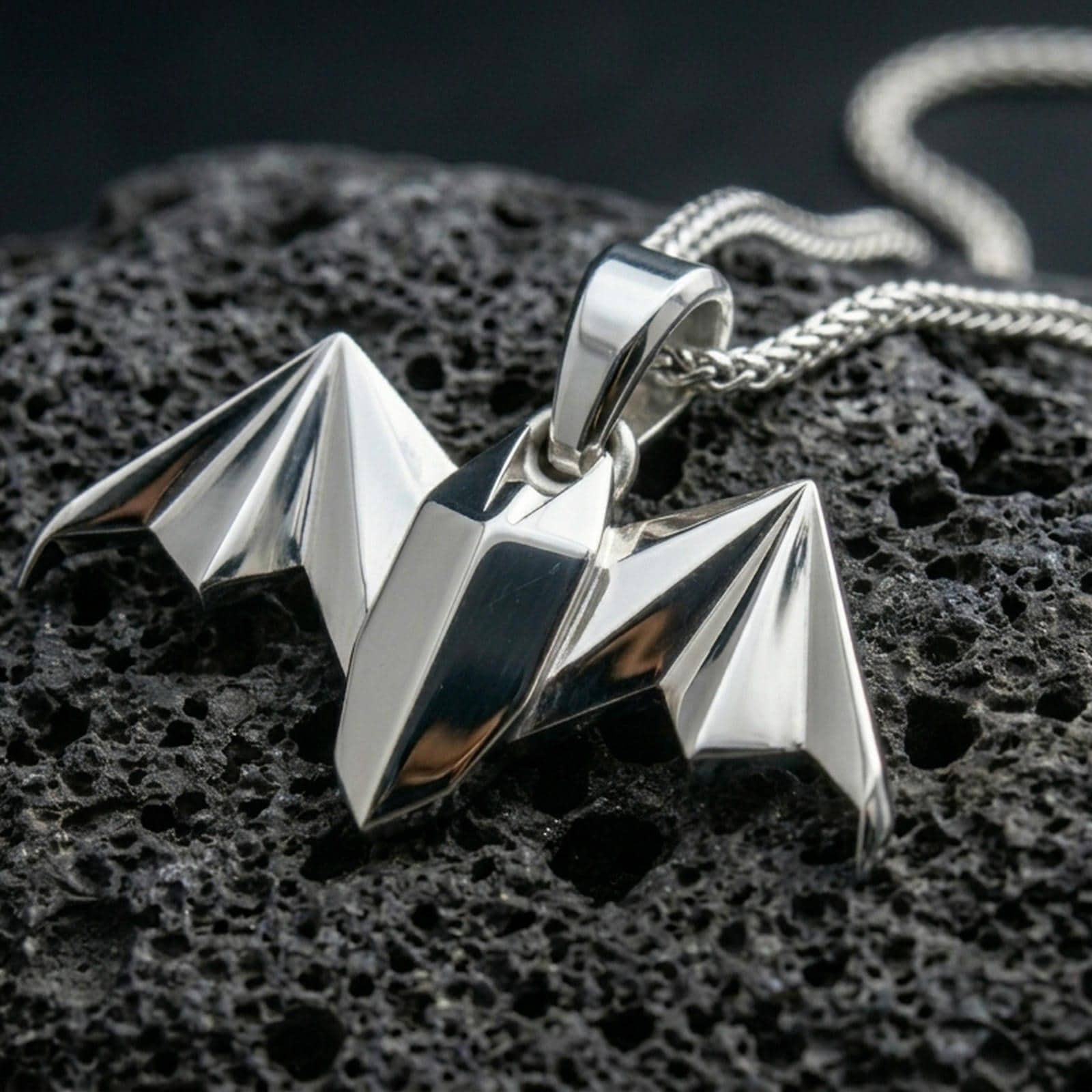 Polished silver bat necklace with angular origami-inspired form, faceted wing structure, and modern gothic animal design placed on textured volcanic rock in a premium studio product shot