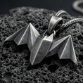 Load image into Gallery viewer, Oxidized origami bat pendant with geometric folded wings and sharp nocturnal silhouette displayed on dark volcanic rock in a close-up luxury jewelry composition

