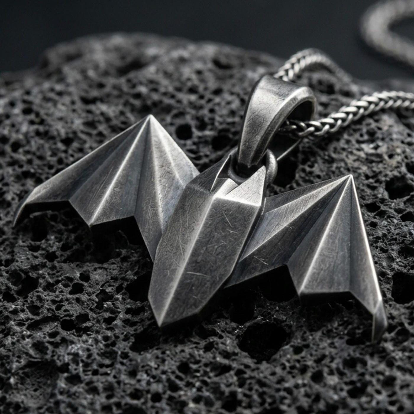 Oxidized origami bat pendant with geometric folded wings and sharp nocturnal silhouette displayed on dark volcanic rock in a close-up luxury jewelry composition