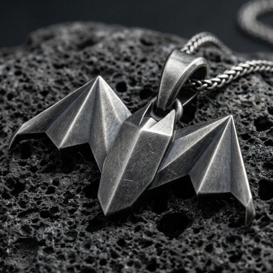 Oxidized origami bat pendant with geometric folded wings and sharp nocturnal silhouette displayed on dark volcanic rock in a close-up luxury jewelry composition