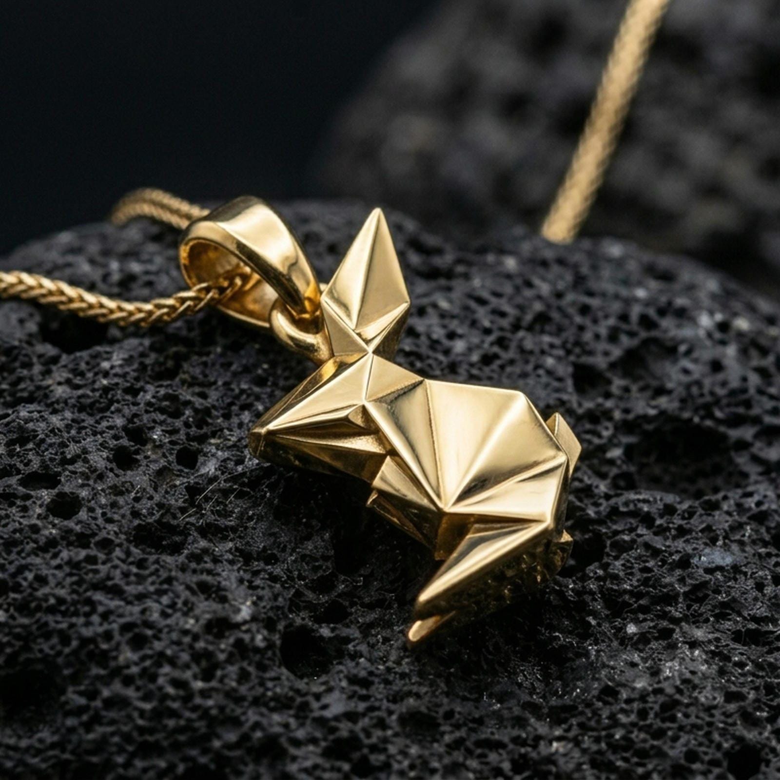Gold plated origami bunny pendant with sharp geometric folds, minimalist rabbit shape, and contemporary sculptural form showcased on black volcanic rock in a refined macro jewelry image