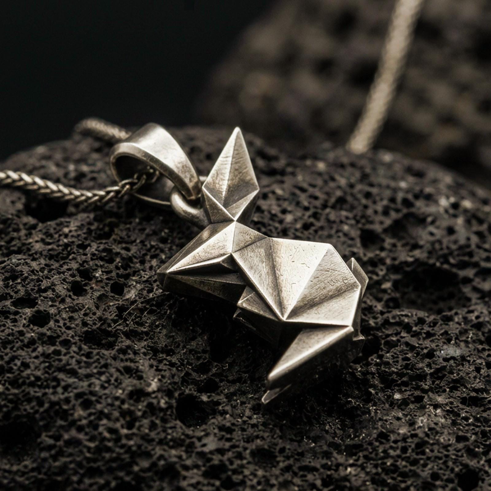 Bronze origami rabbit pendant with geometric folded form and minimalist bunny silhouette displayed on dark volcanic rock in a close-up luxury jewelry composition
