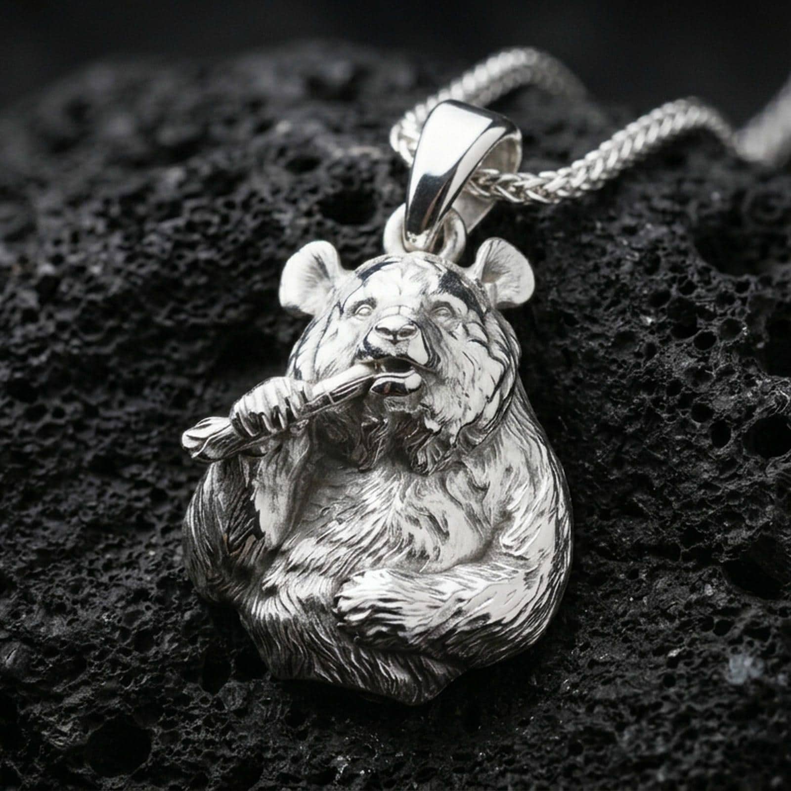 Polished silver panda necklace with sculpted bear face, realistic fur relief, and bamboo detail placed on textured volcanic rock in a premium studio product shot
