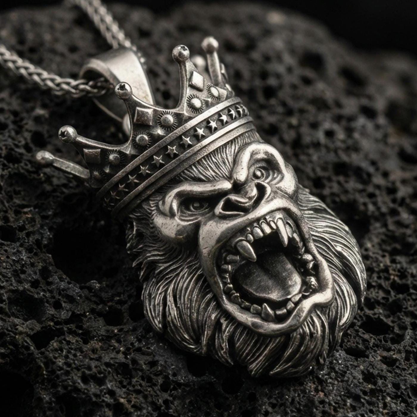 Oxidized gorilla king pendant with roaring ape face, detailed crown, and bold animal head silhouette displayed on dark volcanic rock in a close-up luxury jewelry composition