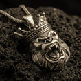 Load image into Gallery viewer, Bronze gorilla king pendant with roaring ape face, detailed crown, and bold animal head silhouette displayed on dark volcanic rock in a close-up luxury jewelry composition
