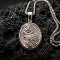 Load image into Gallery viewer, Polished silver owl necklace with intricate feather texture, oval framed design, and elegant wise bird relief placed on textured volcanic rock in a premium studio product shot
