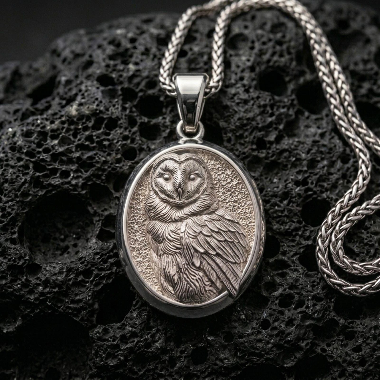 Polished silver owl necklace with intricate feather texture, oval framed design, and elegant wise bird relief placed on textured volcanic rock in a premium studio product shot