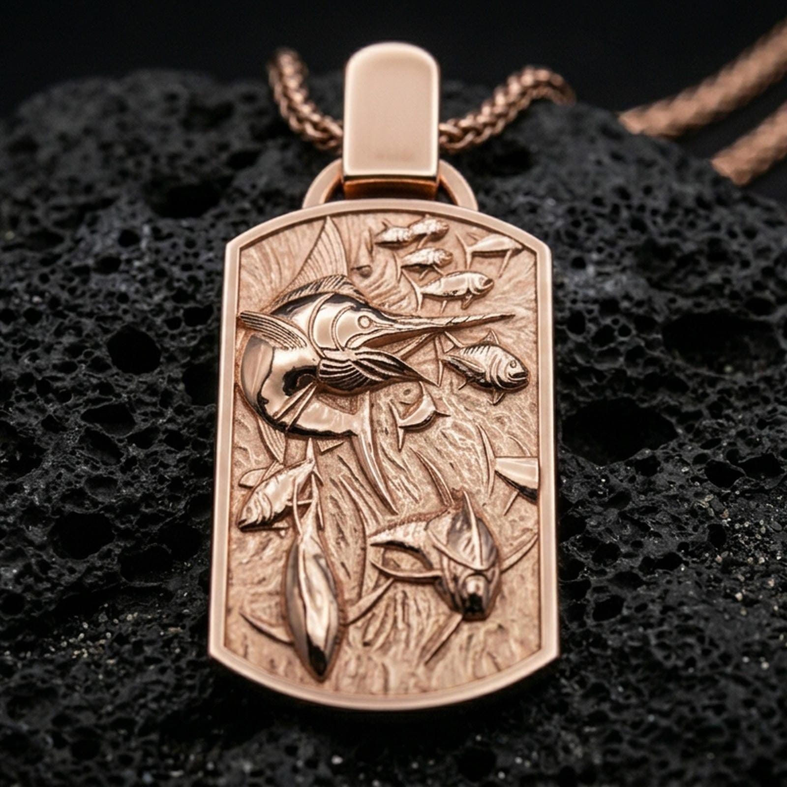 Rose gold plated swordfish necklace with sculpted sea life relief, detailed fish composition, and elegant rectangular medallion form resting on volcanic rock in an elegant close-up product presentation
